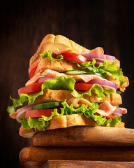 Big size homemade juicy sandwich made of layers with sliced loaf of bread, lettuce, tomato, onion rings, cucumber and ham served on wooden chopping boards against dark brown background for dinner