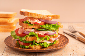 Large homemade sandwich made of layers with sliced loaf of bread, lettuce, tomato, onion rings, cucumber and ham served on brown plate on tiled table at kitchen prepared for dinner as fast food