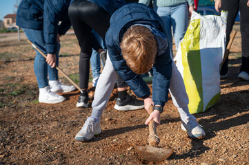 Close-up of a boy bending down to shovel soil, with others assisting in a group tree-planting...