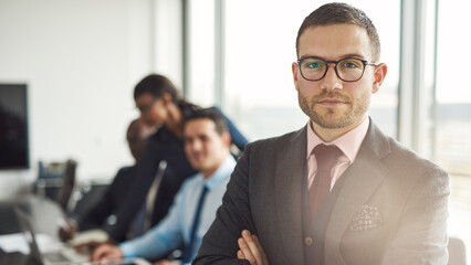 Confident handsome young businessman stands in a boardroom, surrounded by diverse colleagues. He has his arms crossed and looks directly into the camera with a confident smile - 756994551