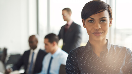 Confident African business woman stands in a boardroom, surrounded by business men. She has her arms crossed and a professional smile on her face. She is in formal clothes and looks into the camera - 756994556