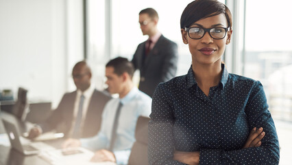 Confident young African business woman stands in a boardroom, surrounded by business men. She has her arms crossed and a professional smile on her face. She is in formal clothes and looks into the cam - 756994565