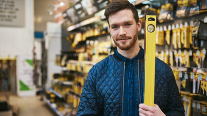 Young smiling handyman with beard carrying a yellow builders or carpenters level in a tool shop - 756995964