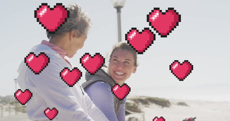 Image of floating red pixel hearts, over happy women talking on beach