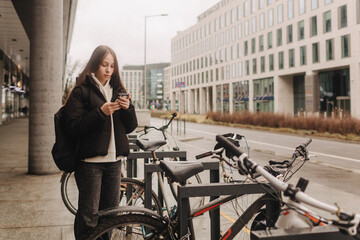Fototapeta na wymiar School age girl renting an electric scooter or bicycle using smartphone, making contactless payment through mobile app. Bicycle rental in the city in the autumn-winter season