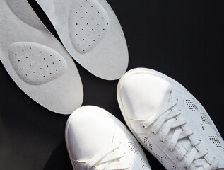 Leather orthopedic insoles and white sports shoes on black background. Foot care accessories. Top view.