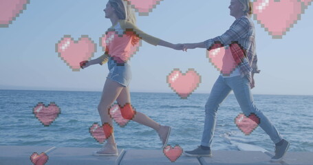 Multiple red heart icons floating against caucasian couple holding hands walking near the sea