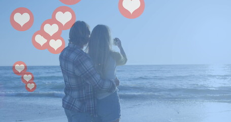 Multiple red heart icons floating against caucasian couple taking picture at the beach