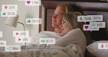 Image of social media notifications over smiling couple relaxing in bed embracing