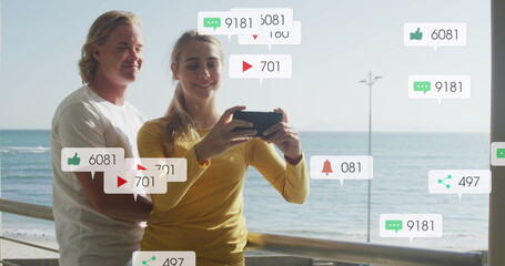Image of social media notifications over smiling couple taking selfie on holiday by the sea