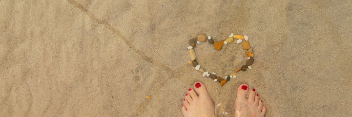 banner of top view of beautiful female legs with red pedicure and heart made of sea pebbles on sea sand.concept of relaxation by the sea on a bright, hot and sunny day on the beach. soft focus