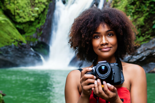 girl photographer. a young cheerful afro girl in a red top stands with a large camera in her hands, a beautiful waterfall from behind, photographer concept