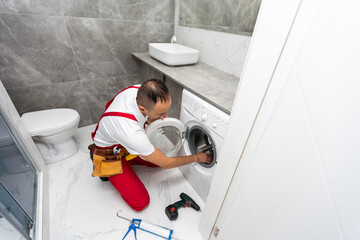 Young smiling happy man plumber handyman in overalls repair fix the washing machine in the kitchen and look to the camera