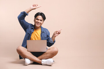 Full length of young Asian man sitting on floor with laptop on his laps, pointing aside to the...
