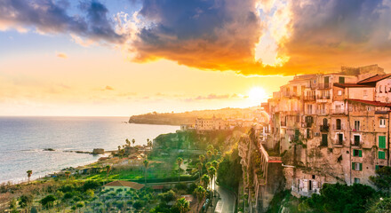 beautiful sunset landscape above sea coastline in Tropea, Italy. Antique buildings on a high rock cliff with green palm trees and scenic cloudy sunset sky on background