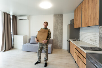 Smiling man in camouflage holding cardboard box in new apartments
