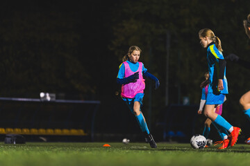 young girls in blue uniforms playing soccer on a grassy field at night, teamwork in sport. High...
