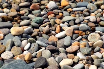 Pebble sea beach with stones of different size colors shimmer in sun in sunny weather. Beautiful stones surface on sea coast. Coastal texture, shoreline, coastline with smoothy rocks.