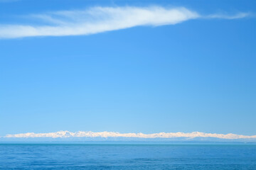 Natural blue background with snowy mountains on sea coast in sunny day with clear sky white clouds. Calm ripples on water surface. Scenic sea horizon and snowy peaks of hills on distance.