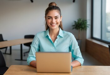 A smiling woman is focused on her laptop in a bright office space. She wears a turquoise shirt.