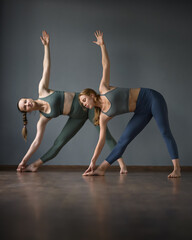 Two women doing yoga. Standing Triangle Pose. utthita trikonasana asana. training in Pilates, stretching in gym. Exercises to stretch leg. Wooden reflective surface. Gray background. Side view.