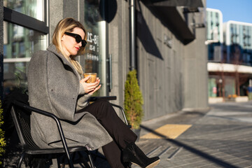 Fototapeta na wymiar Young woman enjoying a coffee, sitting with mobile phone on the cafe terrace on the old city street during a sunny day