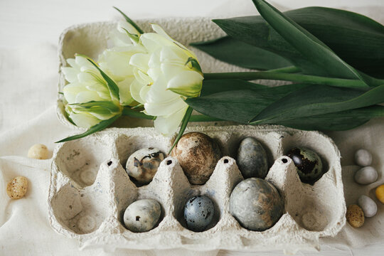 Happy Easter! Stylish easter eggs in tray and tulips on rustic table with linen cloth.  Modern natural dye marble eggs with spring flowers still life. Top view