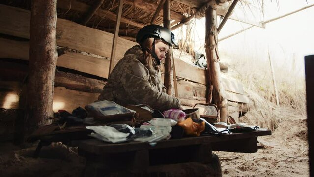 A woman military medic sits inside a bunker, meticulously organizing her medical kit, preparing to provide essential medical services to injured soldiers. Camera 8K RAW. 