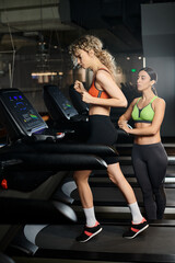 Fototapeta na wymiar good looking sporty female coach and her blonde client in sportwear exercising on treadmill in gym