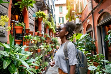 Schilderijen op glas African american girl in casual cloth with backpack travels along a narrow Italian street with lots of green plants. Summer travel concept. © Tanya