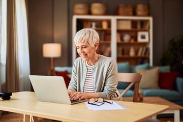 A happy senior adult woman typing on a laptop at a desk while working from home