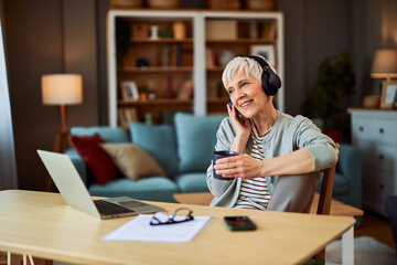 A satisfied senior adult woman listening to music while sitting in front of a laptop and having a...