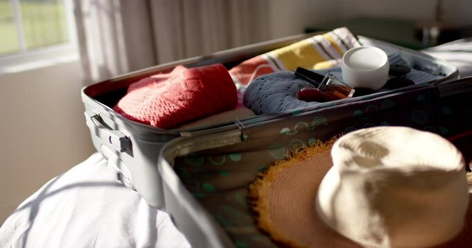 A suitcase lies open on a bed, filled with clothes and a straw hat