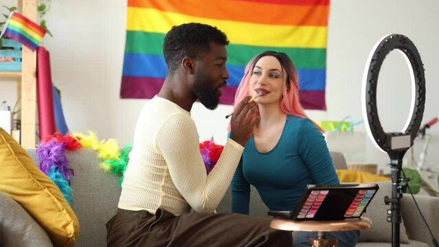 African American friend puts makeup on his friend live for social networks preparing a beauty session before going out to celebrate the pride party with a rainbow flag in the background