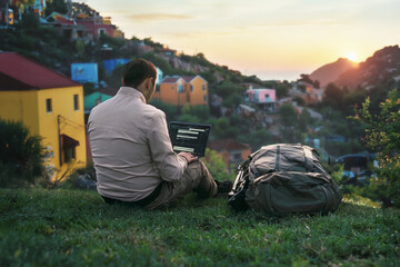 A digital nomad sits on a grassy hill overlooking a scenic village at sunset, working on a laptop...