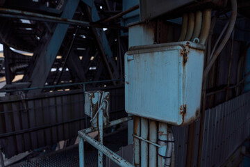 An old electrical box shows signs of wear and rust, part of an intricate network within an industrial framework. The complex steel construction in the background adds depth to the image.