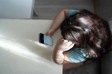 A pensive young girl sits at a table, deeply engrossed in the content on her smartphone. The indoor...