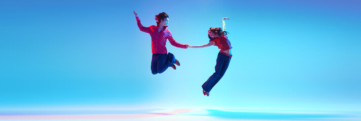 Dynamic image of young couple dancing retro dance, jumping against blue background in neon light. Concept of hobby, dance class, party, 50s, 60s culture, youth. Banner. Empty space to insert text ad