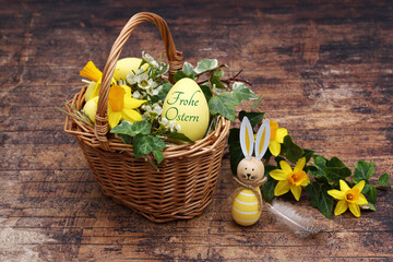 Dekorativer Osterkorb mit Narzissen, Efeu und einem Osterei mit der Aufschrift „Frohe Ostern“.