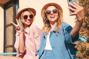 Two young beautiful smiling hipster female in trendy summer clothes. Sexy carefree women posing on street background. Positive models having fun, hugging at sunset. In hat and sunglasses. Take selfie