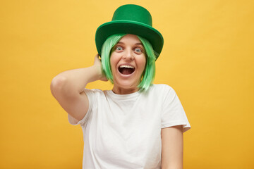 Overjoyed attractive woman with green hair wearing leprechaun hat standing isolated over yellow...