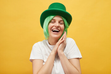 Pleased beautiful woman with green hair wearing leprechaun hat standing isolated over yellow...