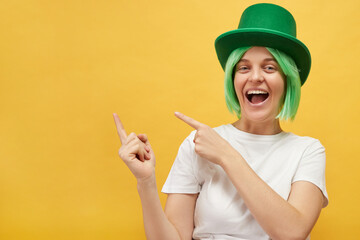 Celebrating St. Patrick's Day traditions. Overjoyed woman with green hair wearing leprechaun hat posing isolated over yellow background pointing at advertisement area copy space