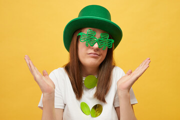 Displeased uncertain woman wearing green hat and clover glasses standing isolated over yellow...