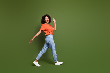 Full length photo of adorable friendly woman wear orange t-shirt waving arm hi walking empty space...