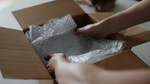 Closeup of unrecognizable man putting breakable fragile glassware in bubble wrap into cardboard box to moving to new house. Male packing fragile things into cartoon box, getting ready for relocation.