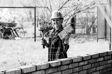 A young female bricklayer in an construction helmet with a construction hammer and a brick in her hands stands and smiles next to a brick wall black white photo - 757180160