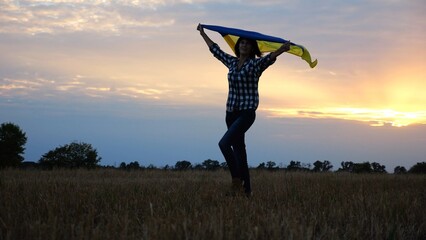 Happy lady walking on barley meadow with a raised over head flag of Ukraine. Ukrainian woman with a lifted blue-yellow banner on a beautiful sunset at background. End of war concept. - 757184110