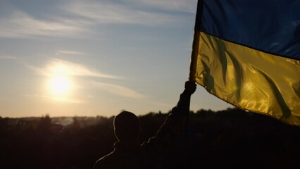 Male soldier stands with raised flag of Ukraine against background of beautiful sunset. Male ukrainian army soldier with a lifted blue-yellow banner in honor of the victory against russian aggression - 757185340