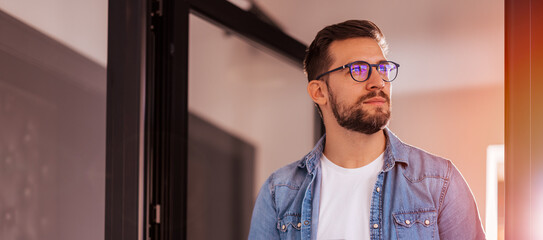 Cheerful entrepreneur in denim shirt, wearing glasses, standing in his office in the morning.